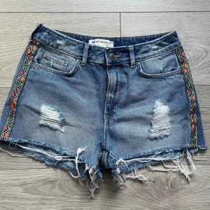 Shorts  - Snygga jeansshorts från H&M Coachella med slitna detaljer och fransar. De har ett coolt mönster längs sidorna i färgglada toner. Perfekta för en avslappnad sommarlook!