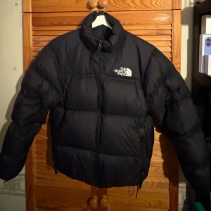 Svart dunjacka från The North Face - Säljer en svart dunjacka från The North Face med dragkedja och hög krage. Perfekt för kalla dagar med sin pufferdesign som håller värmen. Jackan har långa ärmar och logotypen synlig på bröstet och ryggen.