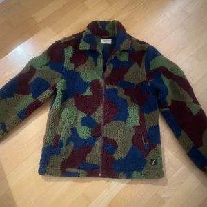 Camouflage teddyjacka från Nudie Jeans - Säljer en cool teddyjacka från Nudie Jeans i ett färgglatt camouflagemönster. Jackan har en dragkedja framtill och är långärmad. Perfekt för att hålla sig varm och stilren under kyligare dagar.
