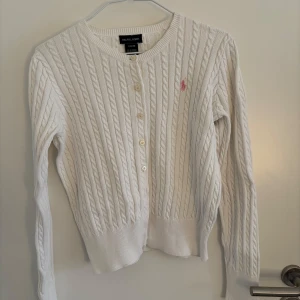 Vit kabelstickad kofta från Ralph Lauren - Säljer en stilren vit kabelstickad kofta från Ralph Lauren med rosa broderad logga på bröstet. Koftan har långa ärmar och knappar framtill. Perfekt för en klassisk look. Säljer för att den är för liten. Det finns en fläck vid höger arm mudd (se bild 5) men syns inte på långt håll!💕 Köptes secondhand men va som ny då. Är som en xs i dam storlek. 
