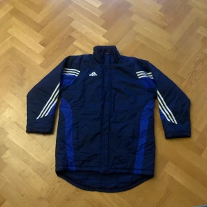 Blå jacka från Adidas - Säljer en blå långärmad jacka från Adidas med vita ränder på ärmarna och logga på bröstet. Jackan har dragkedja och är perfekt för kyligare dagar.