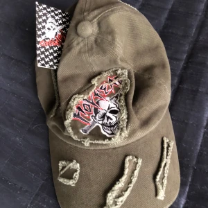 Olivgrön keps med dödskallemotiv - Cool olivgrön keps med ett broderat dödskallemotiv och slitna detaljer. Perfekt för en edgy stil. Märket är okänt men designen är unik och iögonfallande.