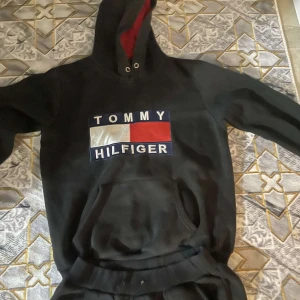 Svart hoodie från Tommy Hilfiger och byxor  - Säljer en svart hoodie från Tommy Hilfiger med det klassiska logotyptrycket i rött, vitt och blått på bröstet. Hoodien har en praktisk magficka och en justerbar huva med snören. Perfekt för en avslappnad stil.