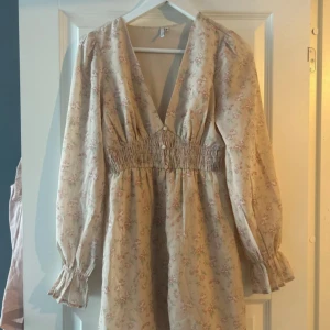 Blommig klänning med v-ringning - Charmig klänning med blommigt mönster i ljusa toner. Den har en v-ringad design och långa ärmar med elastiska manschetter. Midjan är smockad för en bekväm passform.