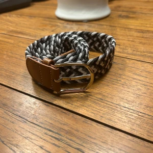 Svart och vit flätat bälte - Snyggt flätat bälte i svart och vitt med bruna skinn-detaljer. Perfekt för att ge en stilren touch till din outfit. Bältet har ett silverfärgat spänne som ger en modern känsla.