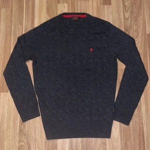 Mörkgrå stickad tröja från Morris 100% merino - Säljer en stilren mörkgrå stickad tröja från Morris med ett diskret rött broderi på bröstet. Tröjan har en klassisk rund halsringning och långa ärmar. Perfekt för kyligare dagar. Trojan är dessutom i 100% merinoull.