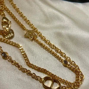 Guldhalsband från Dior - Elegant guldhalsband från Dior med dubbla kedjor och detaljer som en stjärna och en rund berlock med Dior-logga. Perfekt för att ge en lyxig touch till din stil.