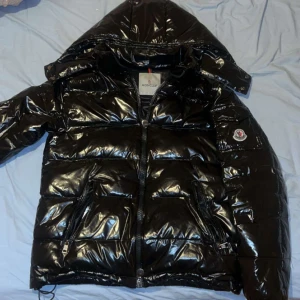 Svart pufferjacka från Moncler - Moncler maya. Storlek 2=S Jackan har en praktisk huva och dragkedja framtill. Perfekt för kyligare dagar med sin vadderade design.luvan går att ta av och finns även scann på jackan skriv för frågor 💗