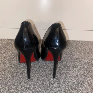 Svarta pumps med röd sula - Eleganta svarta pumps med hög klack och röd sula. Skorna har en spetsig tå och är tillverkade i ett glansigt material som ger en lyxig känsla.