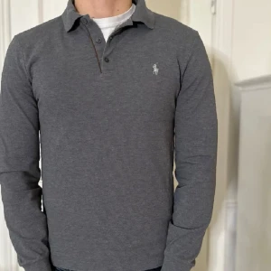 Grå rugbytröja från Ralph Lauren - Snygg grå pikétröja från Ralph Lauren med klassisk krage och knappar. Tröjan har en broderad logga på bröstet och långa ärmar. Perfekt för en stilren look. Ny pris 1299kr