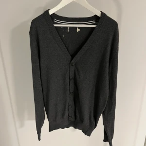 Mörkgrå kofta med knappar - Säljer en stilren mörkgrå kofta med knappar framtill. Den har en v-ringad design och ribbade kanter vid ärmslut och nederkant. Perfekt för lager-på-lager under kyligare dagar. Lappen har råkat slitas av och det har blivit ett litet hål där den satt som man ser på bild 4