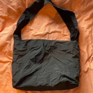 Svart axelväska - Säljer en stilren svart axelväska i syntetmaterial från weekday. Perfekt för att bära en laptop eller andra vardagliga saker. Väskan har en bred axelrem för extra komfort.