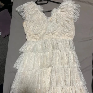 Vit spetsklänning med volanger - Charmig vit klänning i spets med flera lager av volanger. Den har en v-ringad design och är ärmlös, vilket ger en romantisk och luftig känsla. Perfekt för speciella tillfällen som studenten. Säljer min student klänning från by malina storlek xs. Den är precis som ny endast använd en gång. Fläckfri. Skriv för egna bilder.