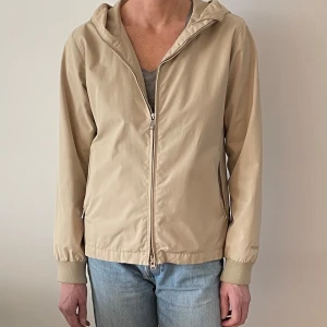 Beige vindjacka från Peak Performance - Snygg beige vintage vindjacka från Peak Performance med huva och dragkedja framtill. Jackan har långa ärmar och ribbade muddar vid ärmslut. Perfekt för vår och höst.