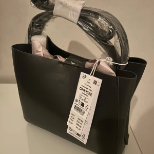 Svart axelväska i syntet - Snygg svart axelväska i syntetmaterial med stilren design. Väskan har en praktisk storlek och kommer med en justerbar axelrem. Perfekt för att bära dina dagliga nödvändigheter med stil.