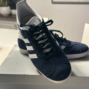 Blå Adidas Gazelle - Snygga blå Adidas sneakers med vita detaljer och snörning. Skorna har en klassisk design med tre vita ränder på sidan och en vit sula. Priset är ej hugget i sten. 