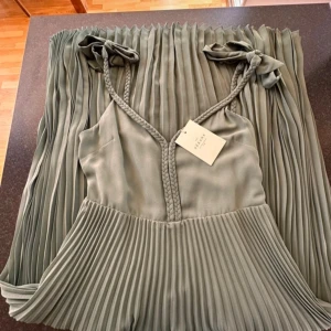 Grön Nella klänning från Sézane - Elegant grön klänning från Sézane med flätade axelband och plisserad kjol. Perfekt för en stilfull look.