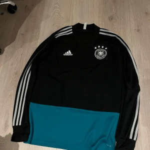 Långärmad Tyskland tröja  - Säljer en Tyskland långärmad tröja från Adidas med vita ränder på ärmarna och ett tryckt emblem på bröstet. Perfekt för sportiga aktiviteter eller som en snygg vardagströja. 