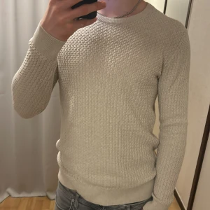 Beige stickad tröja från J.Lindeberg - Säljer en stilren beige stickad tröja från J.Lindeberg, med nypris på 1200. Tröjan har ett snyggt texturerat mönster och ribbade muddar vid ärmslut och nederkant. Perfekt för kyligare dagar.