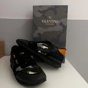 Valentino Garavani sneakers med kamouflagemönster - Snygga Valentino Garavani sneakers i svart med kamouflagemönster och orange detaljer. Skorna har snörning och en modern design som passar perfekt för en trendig look. Storlek 45, syns att de är andvända men fortfarande mycket kvar hör av er vid frågor, kan gå ned i pris vid snabb affär MVH