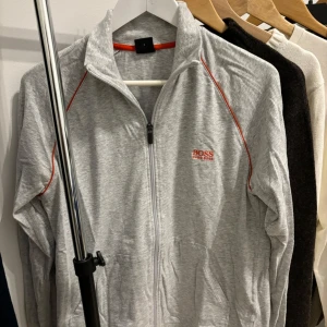Hugo Biss Grå zip-tröja och shorts set  - Snygg grå zip-tröja från Hugo Boss med röd/orange detaljer och logga på bröstet. Väldigt skönt och mjukt material. Tröjan har en dragkedja framtill och är långärmad med matchande shorts. Det är ett set. Nypris: 899kr 