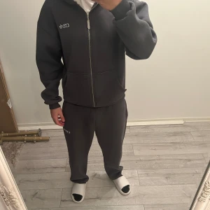 Mörkgrå tracksuit från Curb - Säljer en tracksuit  från Curb med dragkedja och huva. Perfekt för en avslappnad stil. Hoodien har långa ärmar och en bekväm passform.