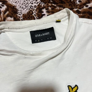 Vit t-shirt från Lyle & Scott - Säljer en klassisk vit t-shirt från Lyle & Scott med korta ärmar och en broderad gul logotyp på bröstet. Perfekt för en stilren och enkel look.