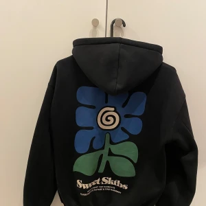 Svart hoodie med blomtryck från Sweet Skibs - Säljer en svart hoodie från Sweet Skibs med ett stort blått och grönt blomtryck på ryggen. Hoodien har en klassisk passform med dragsko i huvan och en stor ficka framtill. Perfekt för en avslappnad stil! 