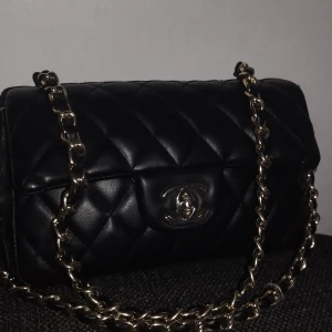 Svart quiltad axelväska med kedja - Snygg svart axelväska i quiltat skinn med en elegant kedja som axelrem. Väskan har en klassisk design med ett metallspänne framtill. Perfekt för att ge en stilren touch till din outfit. 