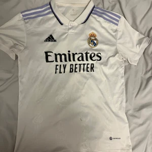 Vit Real Madrid fotbollströja från Adidas - Säljer en vit Real Madrid fotbollströja från Adidas i storlek S, med korta ärmar och klubbens emblem på bröstet. Tröjan har lila detaljer på axlarna och svart text från sponsorn Emirates. 