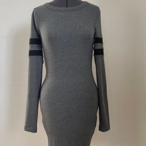 Grå klänning - Grå klänning med lång ärm och svarta ränder på ärmarna. Oanvänd men saknar prislapp.