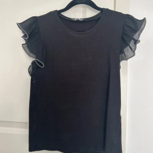 Svart topp med volangärmar från Zara - Snygg svart topp från Zara med volangdetaljer på ärmarna. Tillverkad i ett ribbat material som ger en stilren look. Perfekt för att piffa upp vardagsoutfiten eller för en kväll ute.
