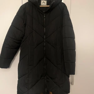 Svart dunjacka från McKinley - Säljer en svart dunjacka från McKinley med huva och dragkedja framtill. Jackan har ett quiltat mönster och långa ärmar, perfekt för kyliga dagar. Den är både stilren och funktionell.