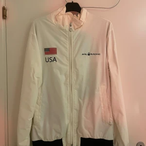 Vit jacka från Sail Racing - Säljer en vit jacka från Sail Racing med USA-flagga och texten 'USA' på framsidan. Baksidan har texten 'UNITED STATES' och en USA-flagga. Jackan har dragkedja och långa ärmar.