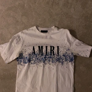 Vit t-shirt från Amiri med blått stänkmönster - Snygg vit t-shirt från Amiri med ett coolt blått stänkmönster över bröstet och ryggen. T-shirten har korta ärmar och en klassisk rund halsringning. Perfekt för en avslappnad stil.