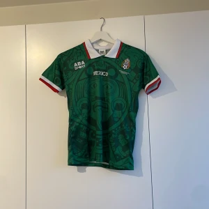 Grön Mexiko fotbollströja från ABA Sport - Säljer en grön Mexiko fotbollströja från ABA Sport med aztekiskt mönster. Tröjan har korta ärmar med röd och vit kant samt en vit krage. Perfekt för fotbollsfans! Retro Mexico hemma ställ 1998 stl 134-140 barn