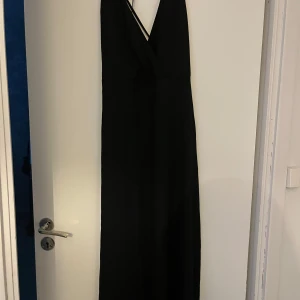 Svart långklänning från DW Wannabe - Elegant svart långklänning från DW Wannabe med v-ringad design och smala axelband. Perfekt för en stilren look.