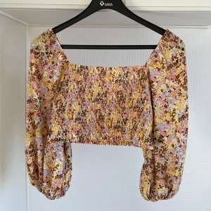 Blommig croppad topp 40 - Säljer en färgglad blommig croppad topp från NA-KD. Toppen har långa ärmar och ett elastiskt smockmönster över bysten. Perfekt för en somrig look!