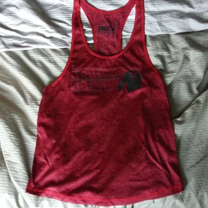 Röd linnetopp från Gorilla Wear - Säljer en röd linnetopp från Gorilla Wear med tryck framtill. Perfekt för träning med sin bekväma passform och stilrena design.