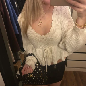 Vit stickad tröja med knytband - Säljer en söt vit stickad tröja med volangdetaljer och knytband i midjan. Tröjan har en v-ringad design med långa ärmar och en ribbad struktur. Perfekt för en mysig dag.