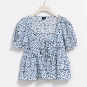 Tie front top Gina - Söt blommig blus i ljusblått med små blommor i mörkblått och vitt. Blusen har korta puffärmar och knytning framtill som ger en fin detalj. Aldrig använd. 