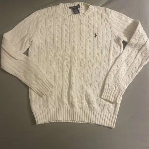 Vit stickad tröja från Ralph Lauren - Säljer en klassisk vit stickad tröja från Ralph Lauren med kabelstickat mönster. Tröjan har långa ärmar och en rund halsringning. Perfekt för kyligare dagar.
