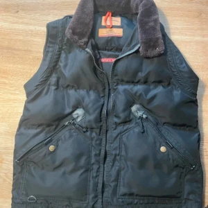 Svart dunväst från Parajumpers - Snygg svart dunväst från Parajumpers med dragkedja och knappar. Västen har en quiltad design och en mjuk krage. Perfekt för kyliga dagar. Finns en lite vit fläck på insidan syn inte när man använde den.
