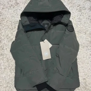 Canada goose macmillan black label  - Säljer en stilren grön dunjacka från Canada Goose med huva och dragkedja. Jackan har en broderad logga på ärmen och är perfekt för kyliga dagar. Den är både varm och bekväm.