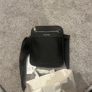 Svart axelväska från Calvin Klein - Snygg svart axelväska från Calvin Klein med ett diskret CK-mönster. Väskan har en dragkedja och justerbar axelrem.  Om pris inte passar ge bud. Helt ny aldrig använd. Nypris 1200kr