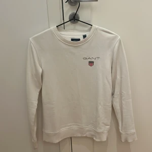Vit tröja från GANT - Säljer en stilren vit tröja från GANT med broderad logotyp och emblem på bröstet. Tröjan har långa ärmar och rund halsringning, perfekt för en avslappnad look.