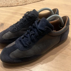  Ralph Lauren Marinblå sneakers  - Snygga marinblå sneakers från Polo Ralph Lauren i mocka och skinn. Skorna har en klassisk design med snörning och diskret logotyp på sidan. Perfekta för en stilren look.