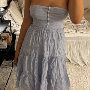 Sötaste sommarklänningen! - Säljer en söt axelbandslös klänning i blå- och vitrandigt mönster. Klänningen har knappar framtill och en dragkedja i ryggen. Perfekt för varma sommardagar! Den har haft ett band till men tog bort det för tyckte den var snyggare utan, hör av er vid frågor🩷