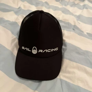 Svart keps från Sail Racing - Snygg svart keps från Sail Racing med vit logga framtill. Kepsen har ett nätmaterial baktill för extra ventilation och justerbar passform. 