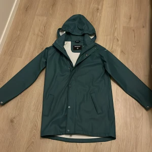 Grön regnjacka från Cross Sportswear - Snygg grön blå regnjacka från Cross Sportswear med huva och dragkedja. Jackan har tryckknappar och är perfekt för regniga dagar. Den är långärmad och har en stilren design.